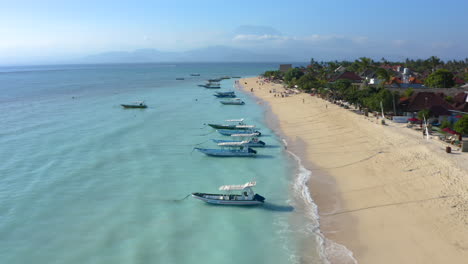 Langsame-Strandaufnahmen-Der-Bali-Insel-Nusa-Lembogan-Mit-Dem-Vulkan-Agung-In-Der-Ferne,-Dramatischen-Tropischen-Momenten-Mit-Segelbooten-Im-Bild,-Kristallklarem-Meer-Und-Gelbem-Sand-Mit-Palmen-Und-Menschen