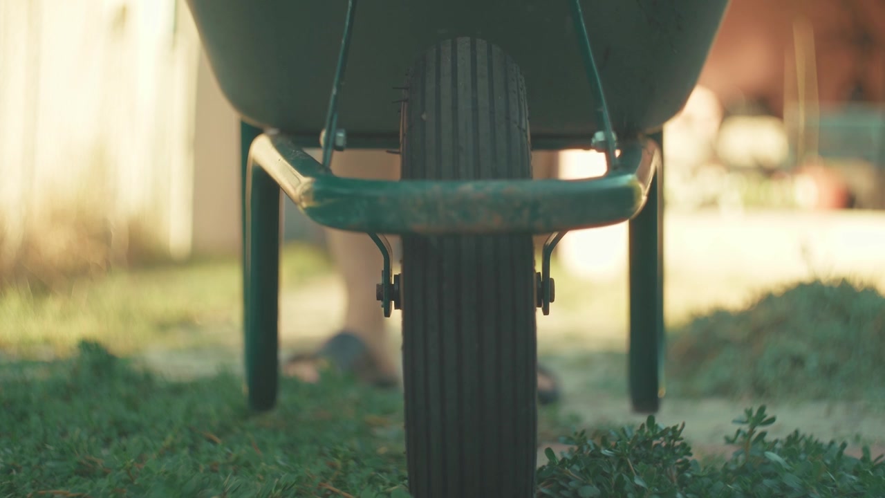 A nostalgic horizontal low angle video shot, moving a garden wheelbarrow from background blur into sharp focus close to the camera on a sunny path at golden hour, using a vintage lens.