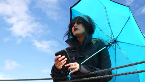 pretty young woman checking the weather or texting on smartphone with umbrella under blue skies as rain storm clouds approach