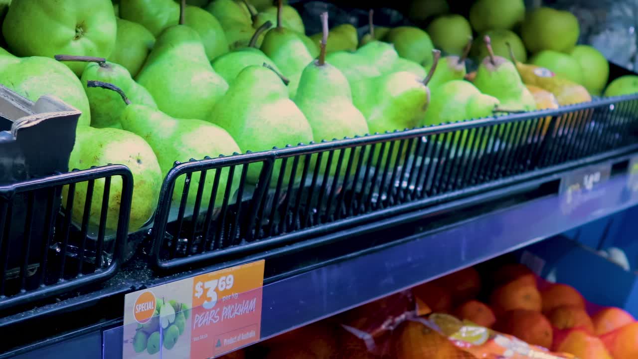 A shopper's hand selects green pears from a display above bags of oranges in a supermarket.