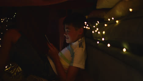 caucasian boy using smartphone while sitting under blanket fort during christmas at home