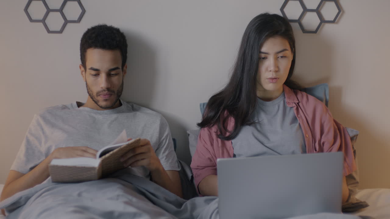 Couple Relaxing in Bed, One Reading, One Working on Laptop