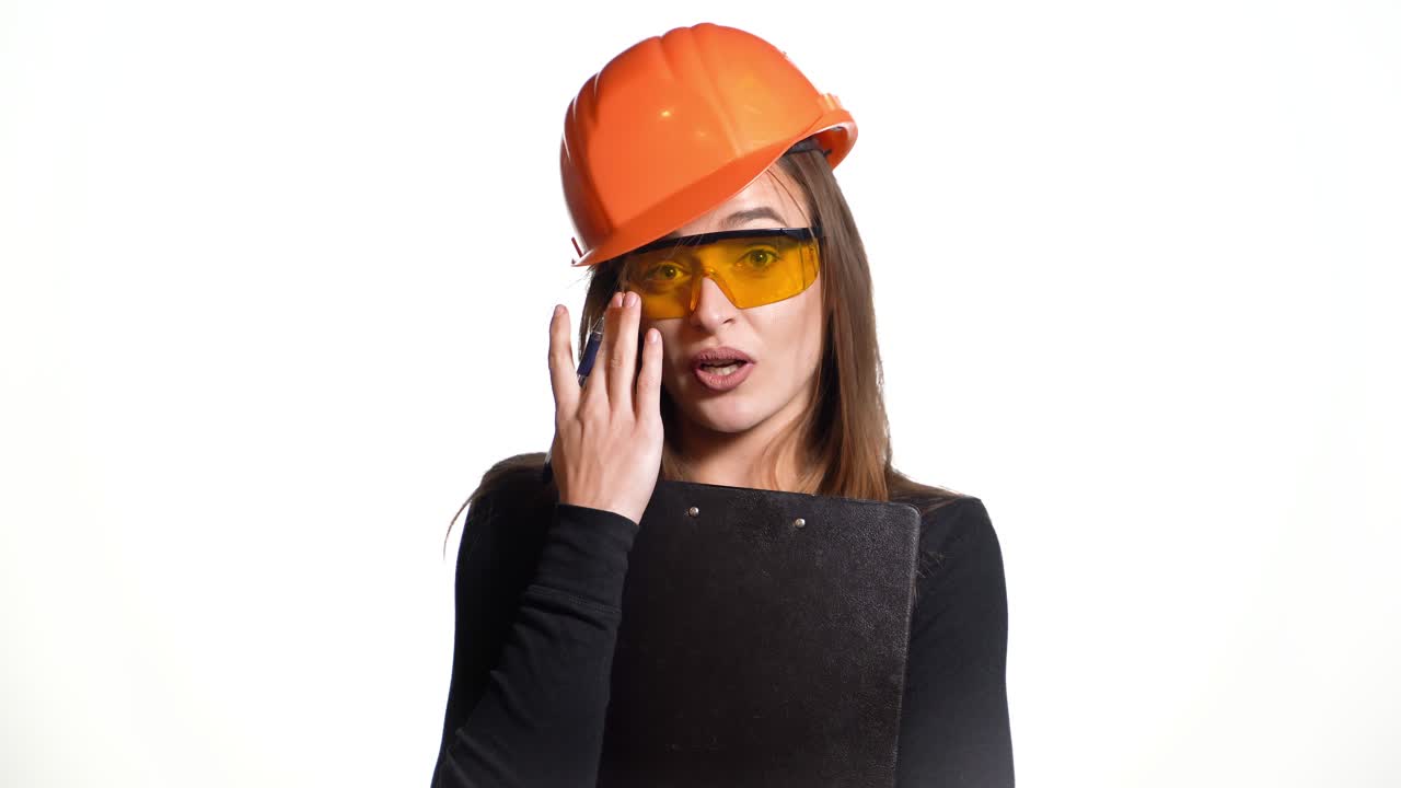 A woman in an orange helmet and protective eyeglasses is talking and holding a pen and folder in her hands on a white background. Features of work of the engineer