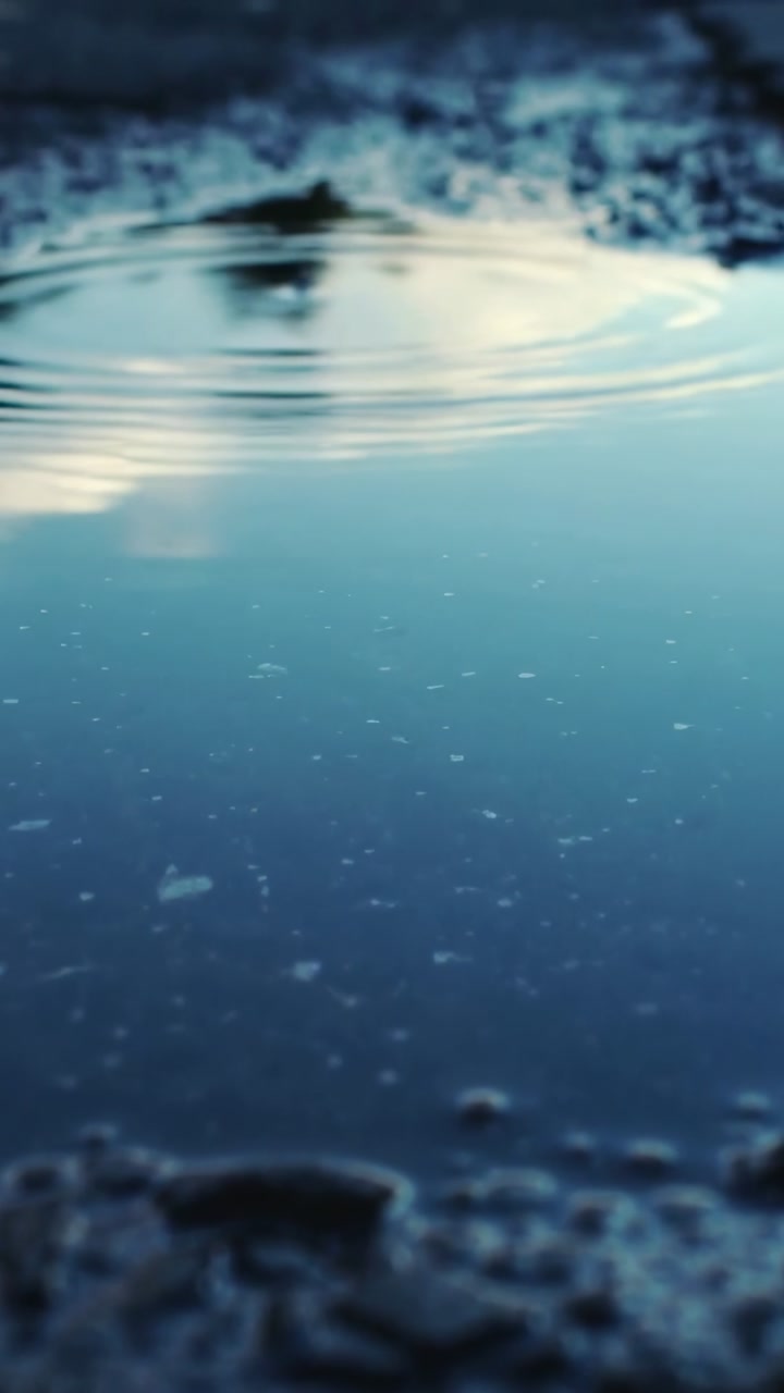 Vertical video: Droplet striking puddle on rocky ground, making round ripples and crown showing sky