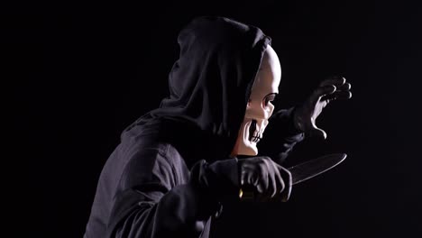 close up side view of scary man in the hooded sweatshirt wearing halloween mask holding a knife and making frightening gesture while running on the black background