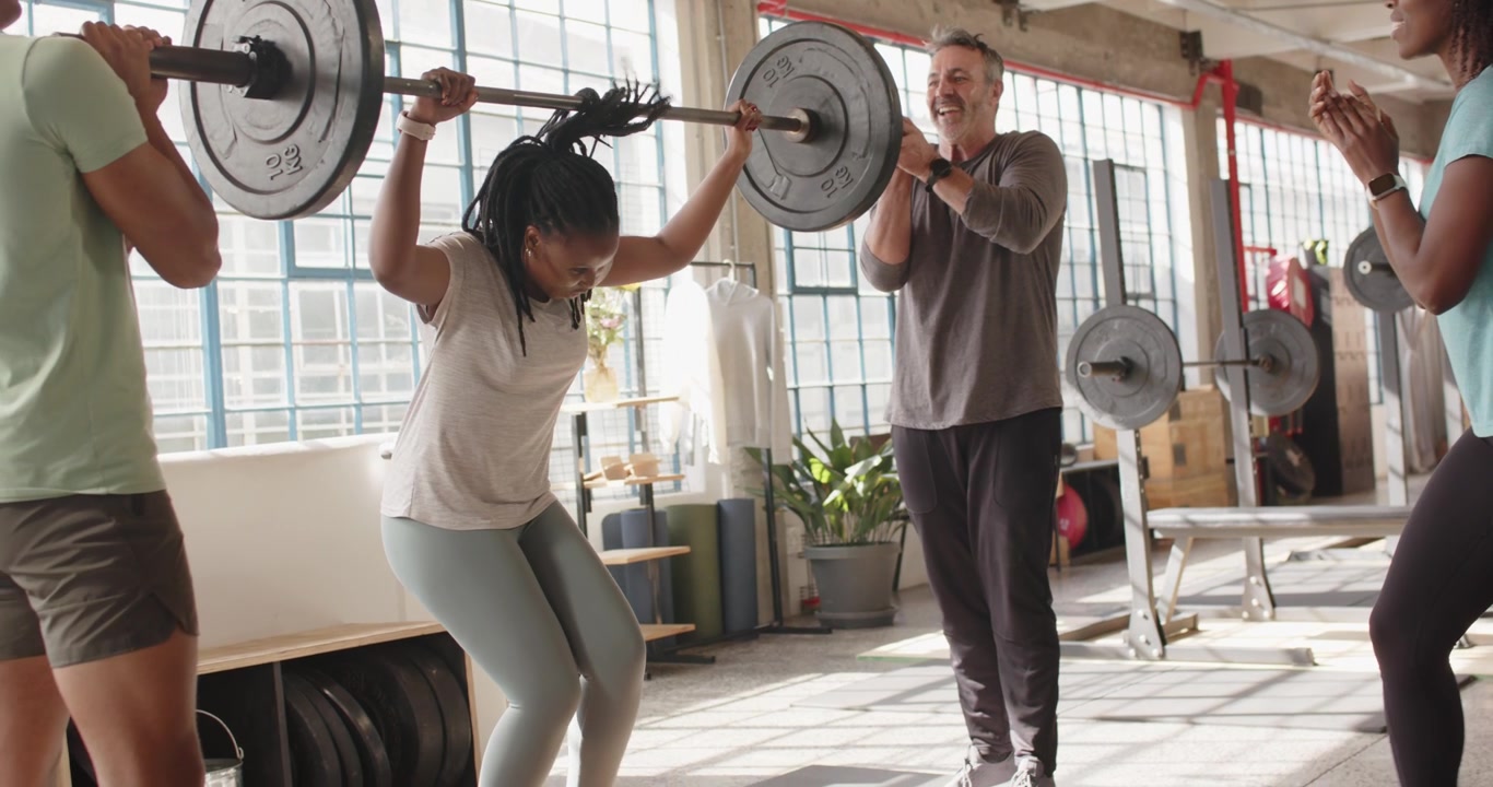 Nhóm đa sắc tộc, huấn luyện viên vỗ tay, người phụ nữ Mỹ gốc Phi đang squat đẩy tạ qua đầu trong phòng gym.