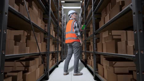 vista trasera completa del cuerpo de un ingeniero masculino asiático con casco de seguridad de pie en el almacén con estantes llenos de bienes de entrega. zoom en pantalla verde teléfono inteligente y mirando alrededor del almacenamiento