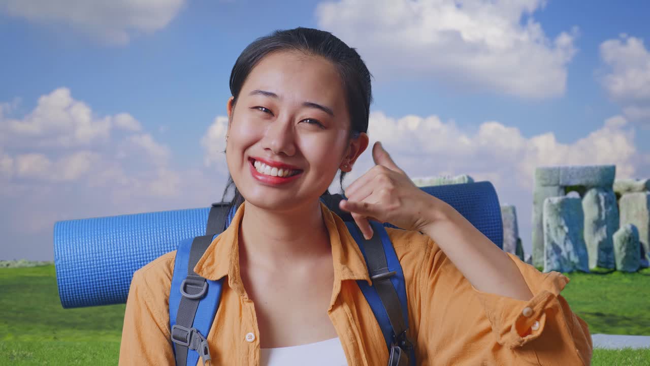 Close Up Of Asian Female Hiker With Mountaineering Backpack Smiling And Showing Call Me Gesture While Traveling In Stonehenge