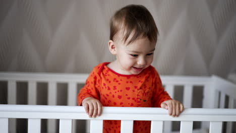 adorable smiling one year old boy stands in his crib laughing cheerfully. lovely baby looks down holding by the handrail.