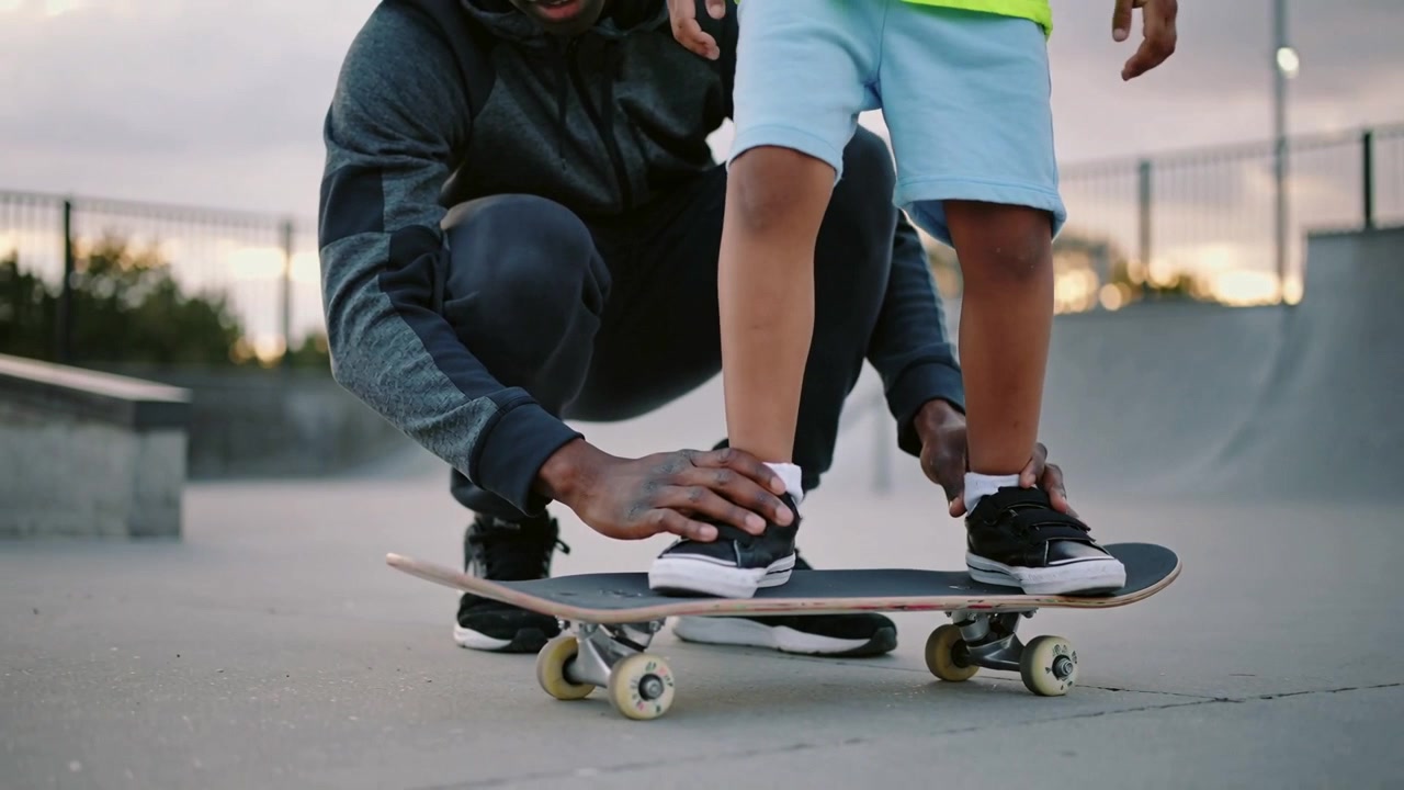 Placing board, kneeling coach steadying child learning balance on skateboard at park in lime shirt