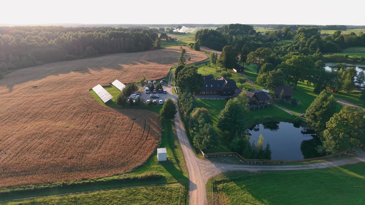 Video con drone che mostra una tranquilla fattoria di campagna delimitata da campi dorati, uno stagno sereno, una foresta lussureggiante ed edifici agricoli bagnati dalla luce del sole del tardo pomeriggio e un ampio movimento aereo