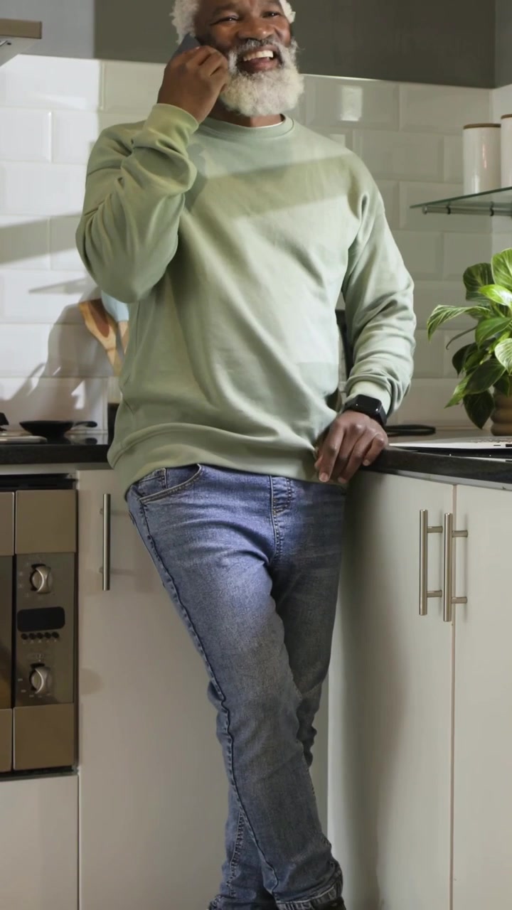 Vertical video: Answering phone, mature man in green top in kitchen checking watch, tapping tablet