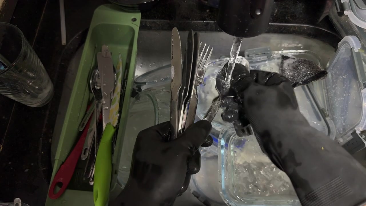 Hands in black rubber gloves scrub and rinse dirty cutlery and glass containers in a cluttered kitchen sink, useful for cleaning, chores, or home lifestyle themes.
