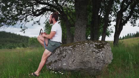 Farmer-With-Scythe-Walks-Toward-Huge-Boulder-To-Sit-And-Take-A-Break-From-Farm-Work