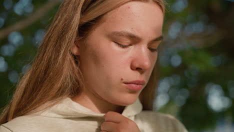 close-up of young woman with blond hair in deep thought, gently touching her hood collar, face softly illuminated by sunlight filtering through tree leaves