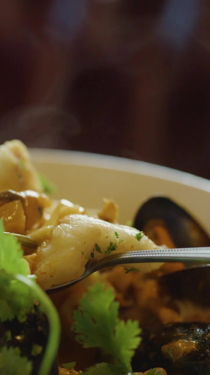 Primo piano di una zuppa di pesce fumante con cozze, calamari ed erbe aromatiche, mentre una mano usa un cucchiaio per servire e mangiare. Illuminazione calda e naturale.