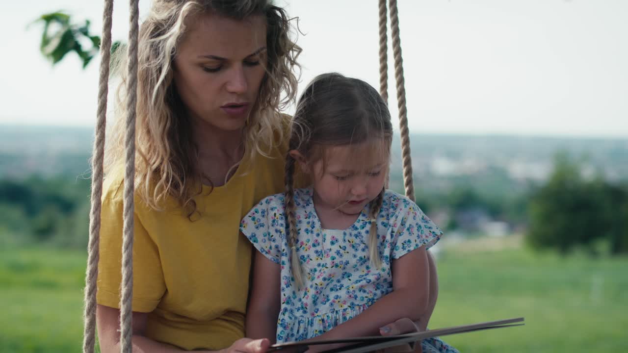 Mom with little girl swinging and reading book in summer day.