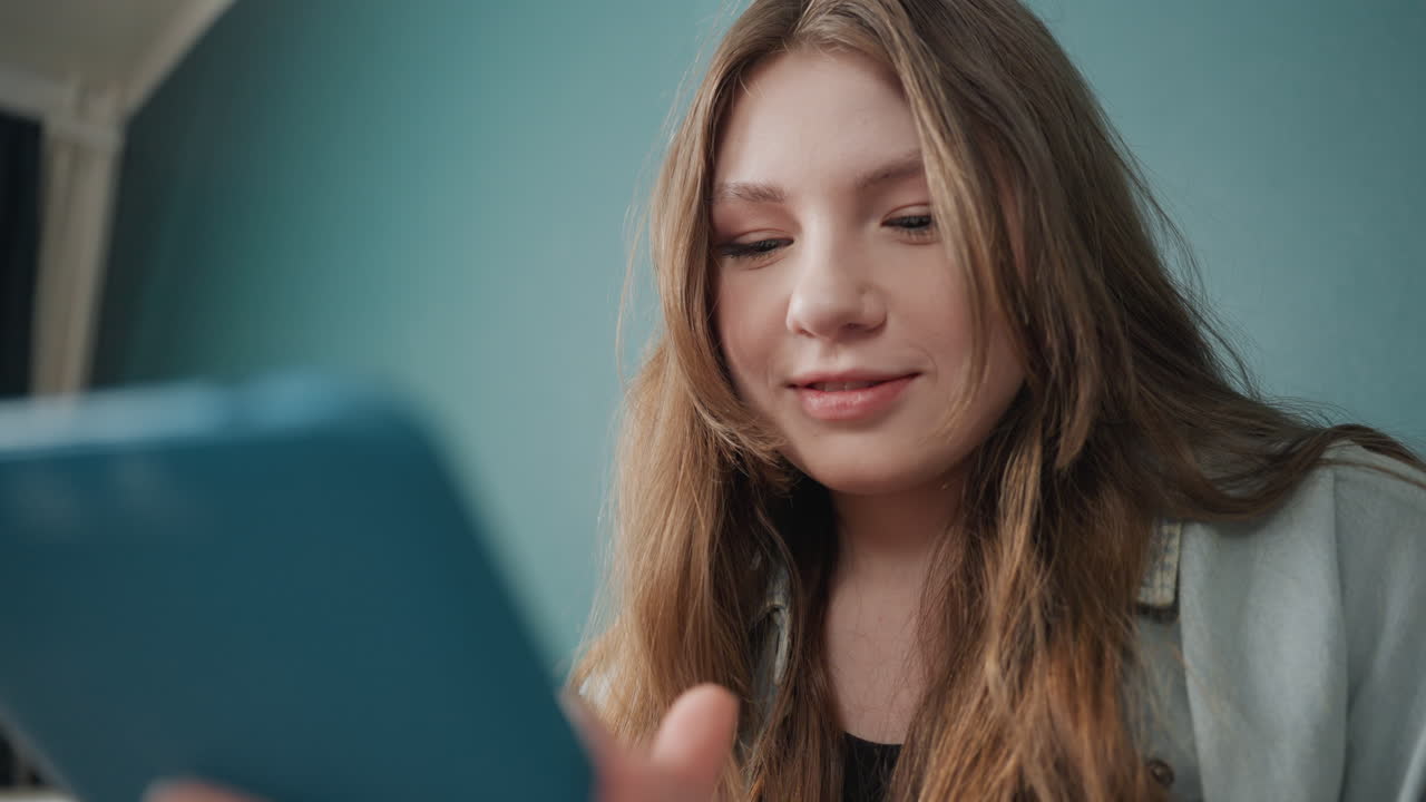 Giovane donna che sorride al tablet in camera da letto, spettatrice coinvolta che si gode i contenuti con il caldo bagliore di una lampada, sfondo di parete color verde acqua, capelli sciolti e maglione, reazione ravvicinata e sorriso gentile mentre scorre un'app social.
