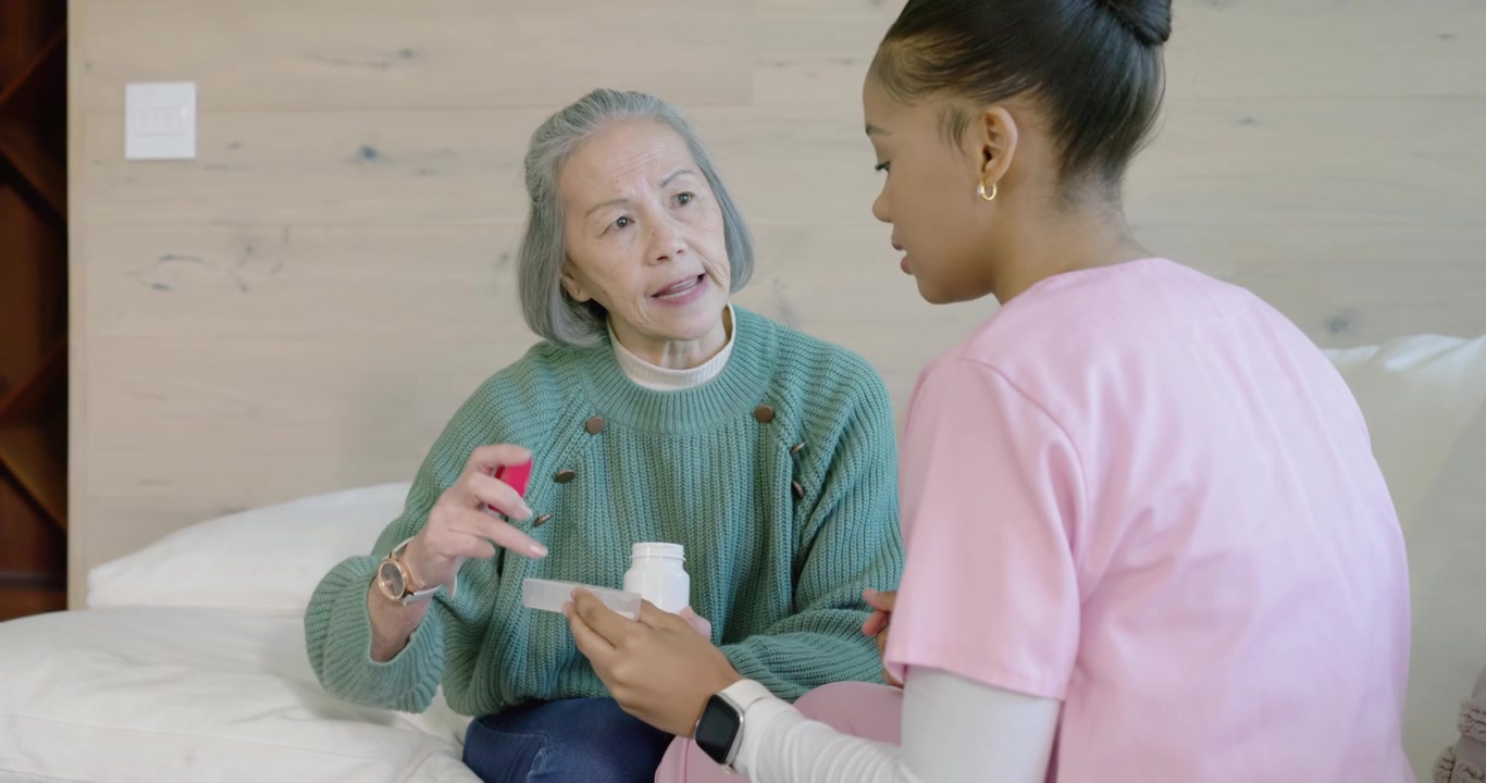 Aidante diverse guidant une personne âgée sur le lit, la personne âgée ouvrant une bouteille à bouchon rouge et versant des médicaments dans une tasse.