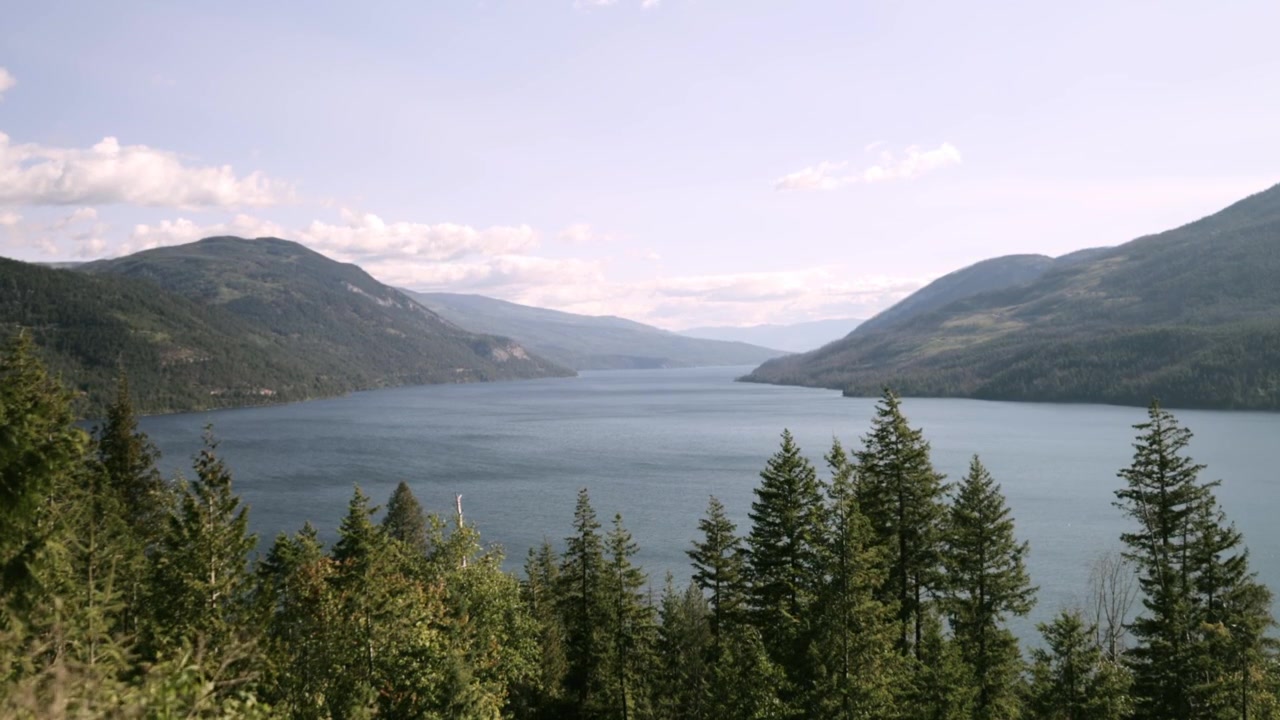Et udsigtspunkt på en bjergside over Adams Lake, der viser dybblåt vand mellem skovklædte højdedrag under et tyndt slør af skovbrandsrøg – stabilt håndholdt perspektiv med blødt dagslys.