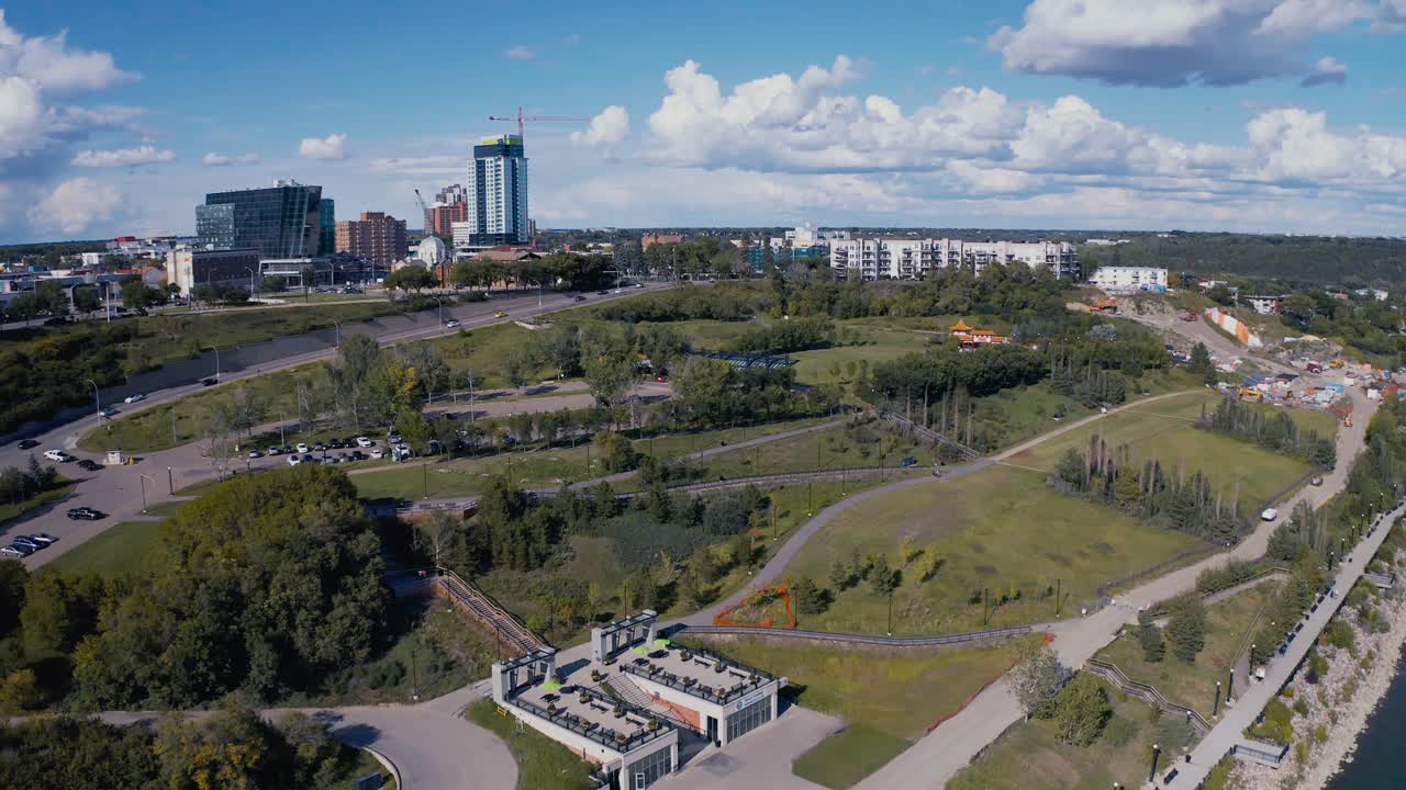 Aerial flyover the dark blue North Saskatchewan River, flyby the Louise Mckinney Public lush Park Chinese Garden Pavilion and oval track birds eye view on a hot summer day with cumulus cloud coverage