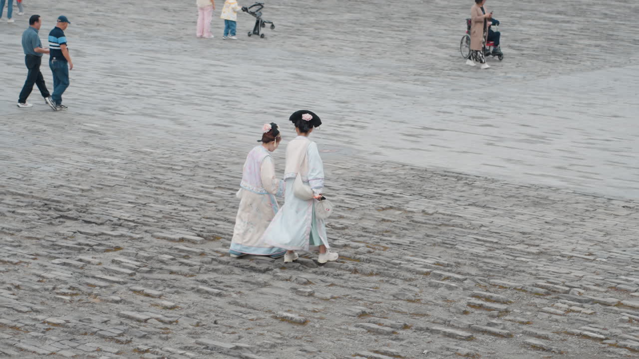 Two women in traditional dresses walking along sandy beach during cultural festival, pastel tones and overcast light, distant crowd with stroller, candid promenade showing quiet companionship