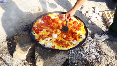 mężczyzna, który robi omlet z omletem z szafranowych pomidorów na patelni na ogniu z drewna w przyrodzie na zewnątrz