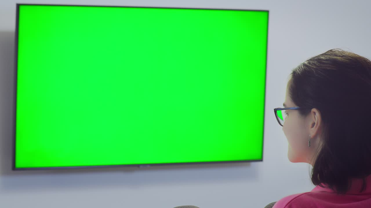 chica con gafas viendo la tv sentada frente a la tv, cambiando de canal con el control remoto de vista trasera, llave de croma