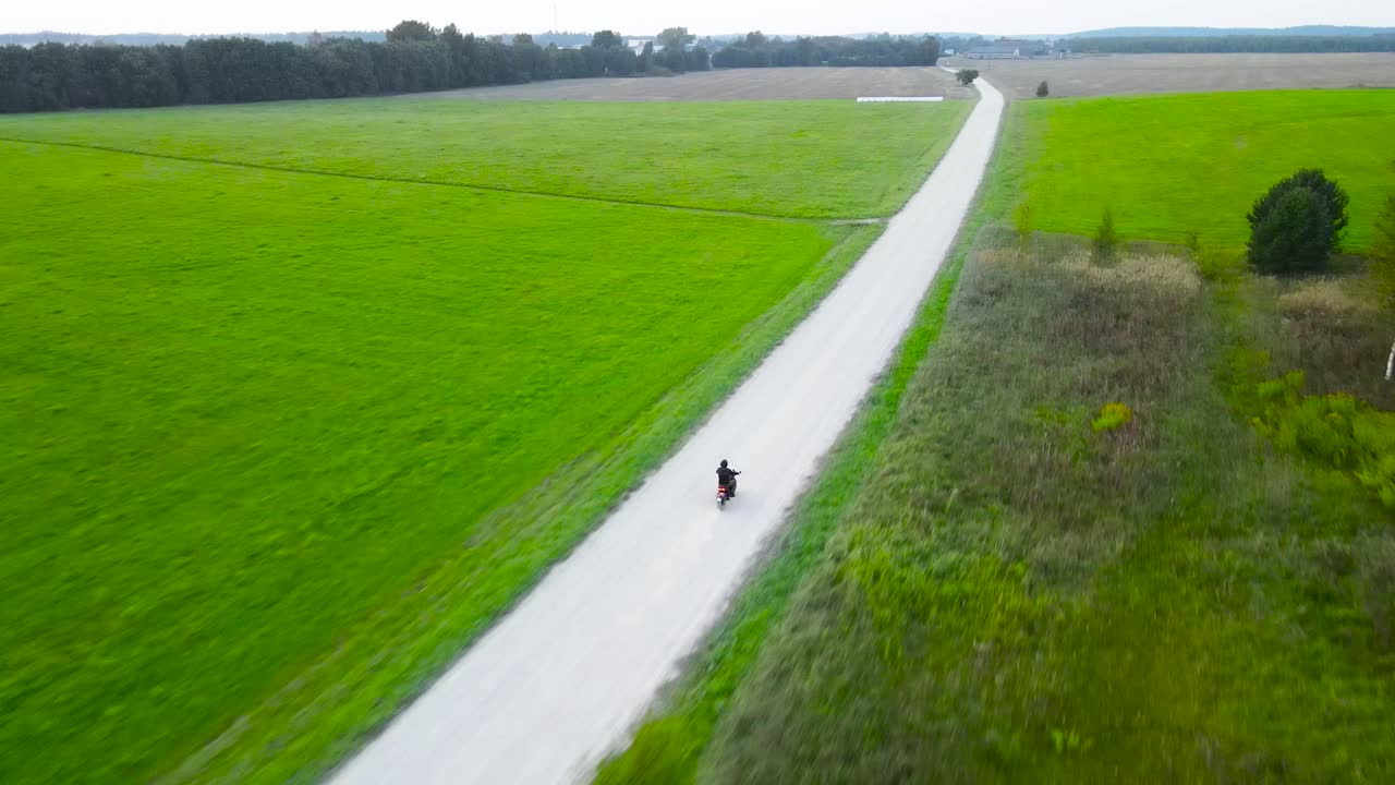 Aerial drone footage following and flying behind a motorcycle driver or person driving on a summer gravel road that is creating dust while the person is driving between green desolate rural fields.