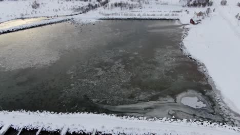 Drone-view-in-Tromso-area-in-winter-flying-over-a-snowy-landscape-surrounded-by-the-sea-and-a-frozen-port-with-boats-in-Norway