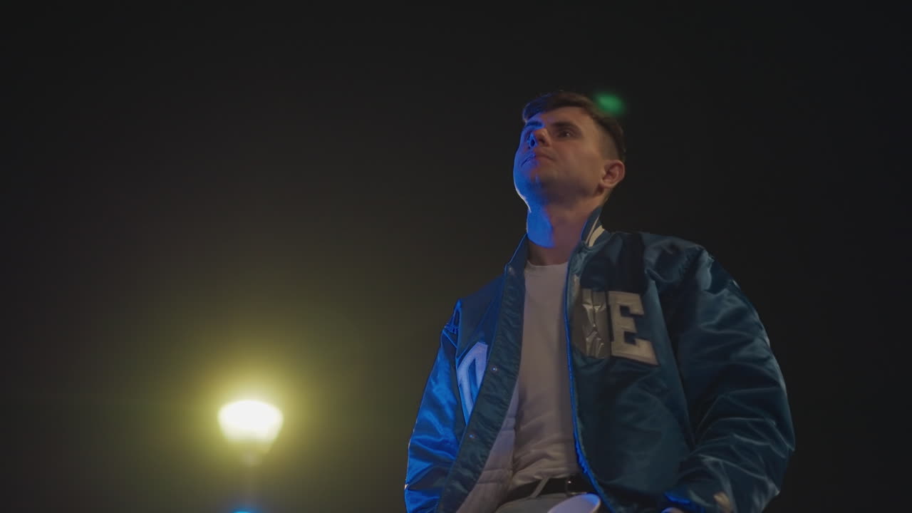 hombre blanco joven bajo la luz de una farola, apoyado en un coche aparcado en un lote con luces de neón, con una mirada contemplativa hacia el cielo, un cigarrillo en la mano, grano cinematográfico melancólico, soledad urbana