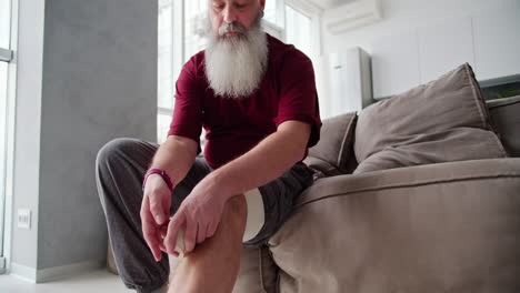 an elderly man with intact hair and a lush beard in a red t-shirt bandages his leg and knee with a bandage while sitting on a brown sofa in a modern apartment