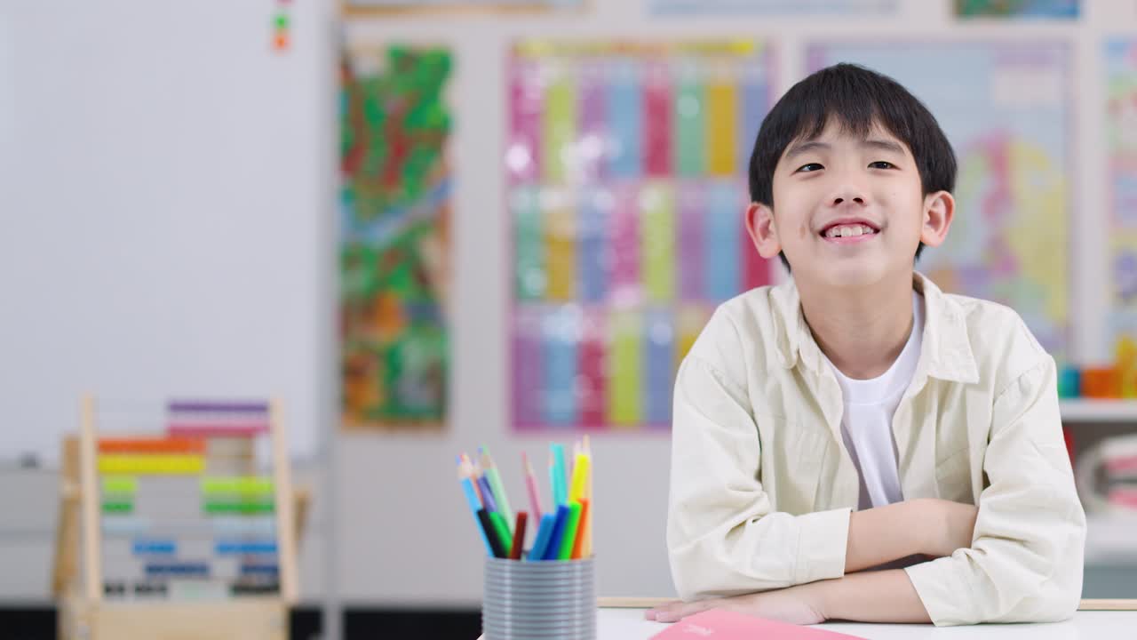 Smiling boy with arms crossed at desk in bright, colorful classroom, steady camera, natural light