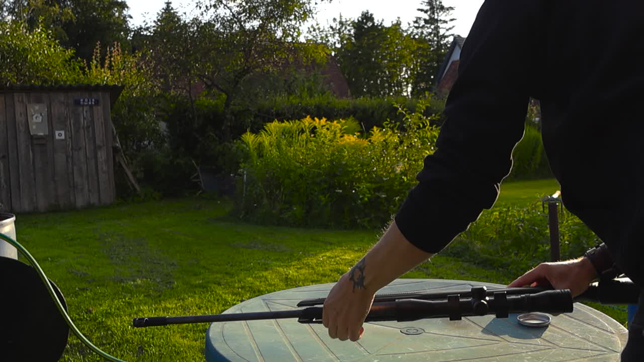 Man with black clothing and tattoos shooting a firearm gun rifle that has a scope in a sunset garden during summer or autumn while sun is creating lense and light flare in the background. Man aims