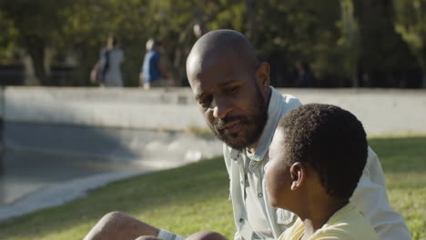 Closeup-of-father-and-son-sitting-on-lawn-in-park-and-talking.