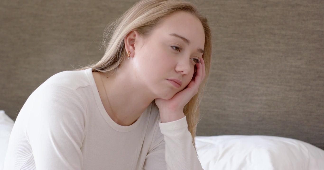Female sitting on bed edge, leaning forward, resting head on hand, reflecting near pink phone