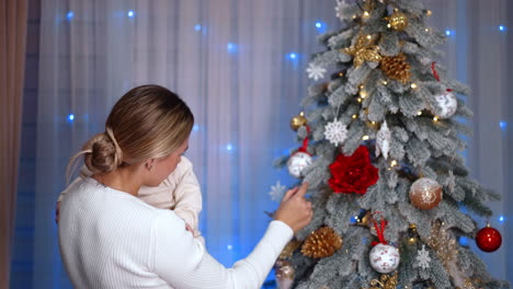 Mother shows the beautifully decorated Christmas tree to her little infant baby. Mom and son celebrate holidays together.