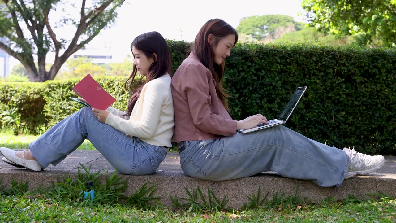 To kvindelige studerende sidder ryg mod ryg udendørs og bruger en bærbar computer og en notesbog til studier.