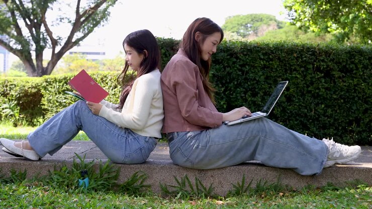 Due studentesse sono sedute schiena contro schiena all'aperto, usando un laptop e un quaderno per studiare.