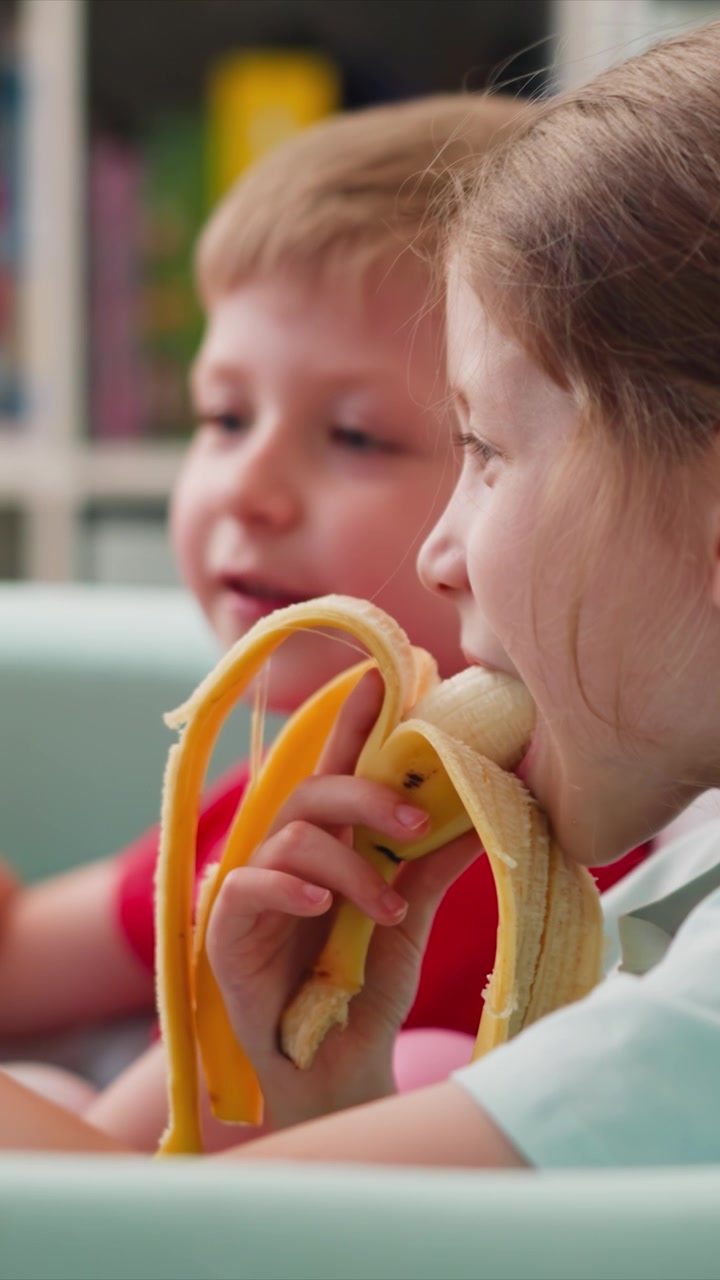 Primo piano di due bambini che mangiano banane insieme. Un bambino tiene una banana parzialmente sbucciata mentre l'altro allunga la mano per prendere il proprio spuntino, entrambi seduti su seggioloni con illuminazione soffusa e in interni.