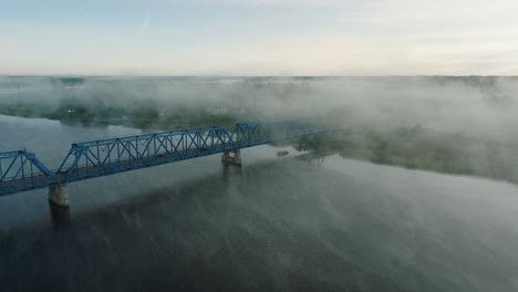 Luftaufnahme-Der-Stahlbrücke-über-Den-Fluss-Lielupe-An-Einem-Sonnigen-Sommermorgen,-Nebel-Steigt-über-Dem-Fluss,-Fahrende-Autos,-Breite-Drohnenaufnahme,-Die-Sich-Vorwärts-Bewegt,-Nach-Unten-Geneigt