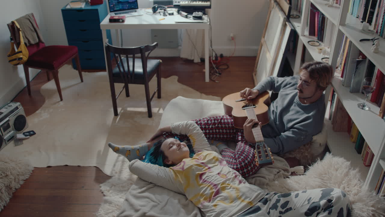 Boy Playing the Guitar to Girlfriend Relaxing on Bed at Home