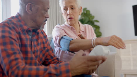 una pareja de ancianos de dos razas sentada en el sofá y comprobando la presión arterial en casa, en cámara lenta.