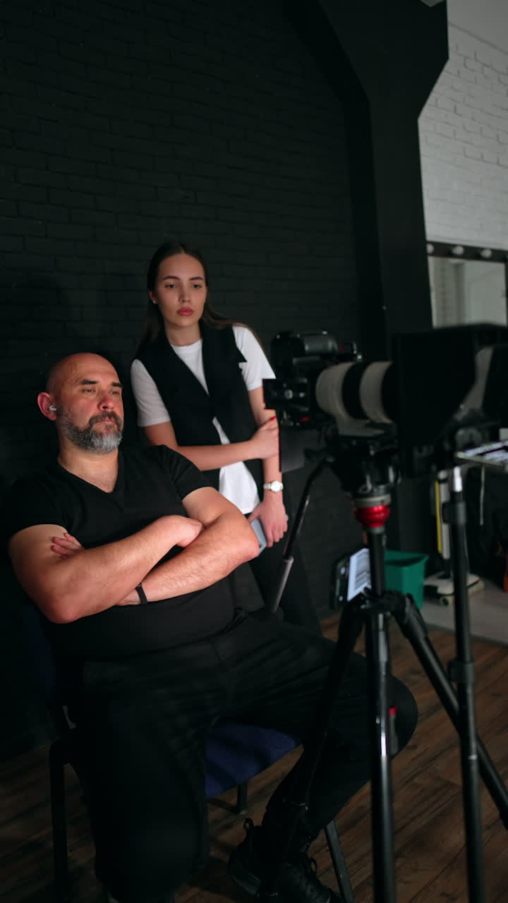 Relaxed Caucasian bearded cameraman sits his hands crossed on his chest. Photographer looks at camera, points at display and addresses the lady standing behind. Vertical video