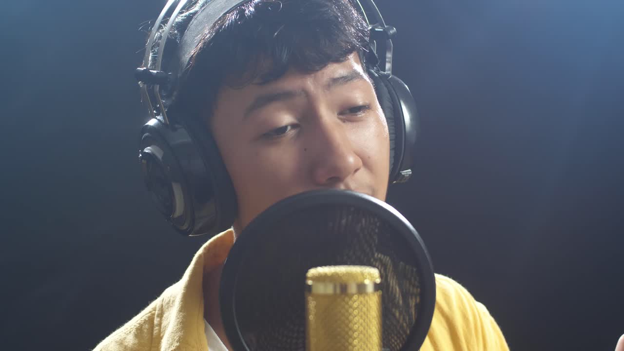 젊은 아시아 소년의 근접 사진. 헤드폰을 들고 검은색 배경의 콘덴서 마이크로 노래하고 있습니다.