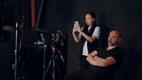 male photographer sits behind the camera on tripod. long-haired woman takes video on her phone. work in modern photo studio.
