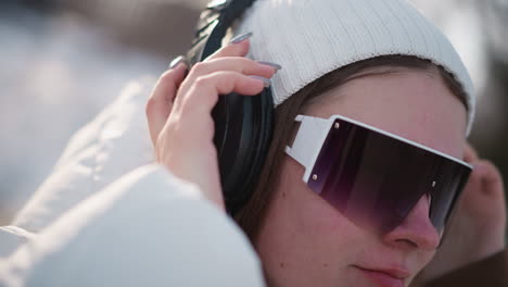 close up of lady wearing over ear headphones smiling while listening to music against winter blur background snow glistening under sun breeze on coat and beanie capturing joyful vibe