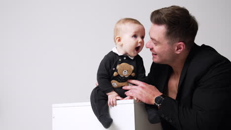 lovely little child sits on the cube. father in black jacket holds the baby and talks to him. white backdrop.