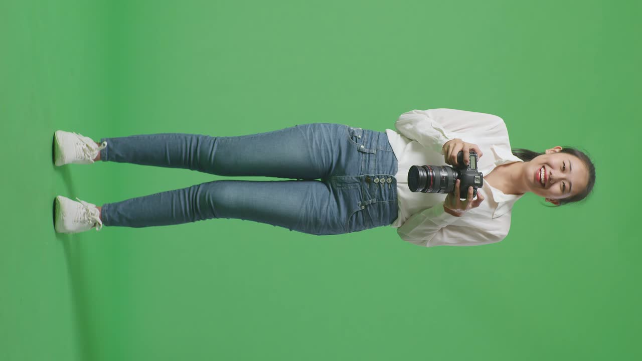 corpo pieno di fotografo asiatico guardando le immagini nella telecamera e sorridendo essendo soddisfatto del risultato mentre si trova sullo sfondo dello schermo verde nello studio