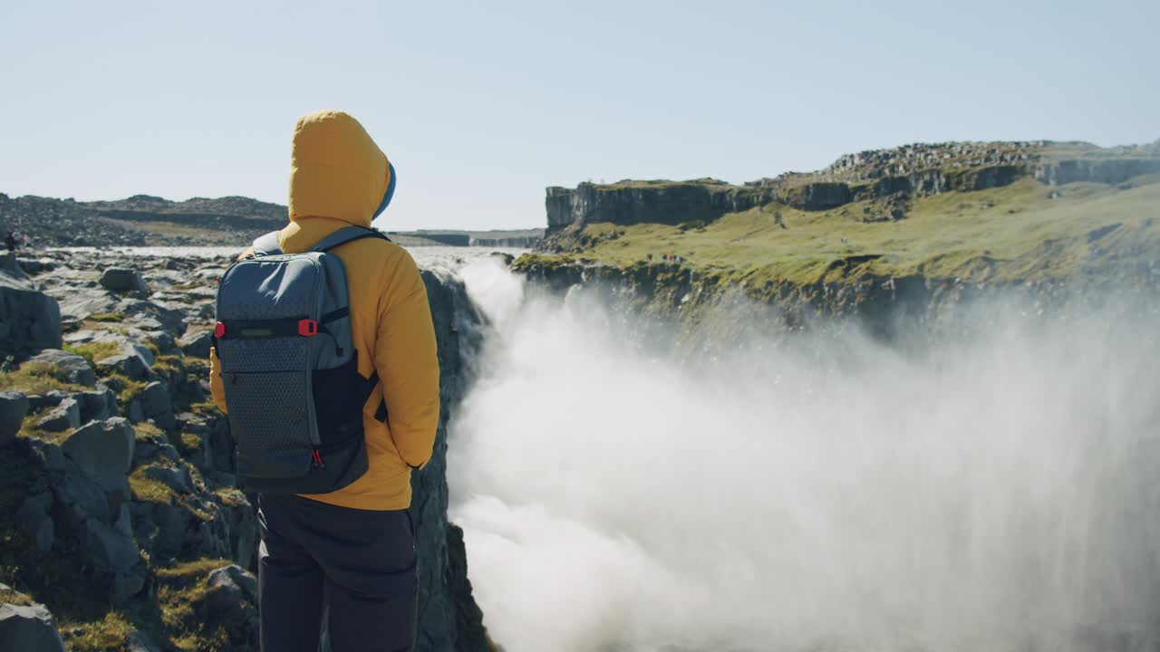 vista trasera de un hombre con chaqueta amarilla y mochila de pie en el borde de un acantilado mirando y disfrutando de la cascada de detifoss en islandia